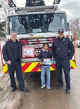 Two firefighters and a young girl standing in front of a fire truck, holding a couple prizes and a framed picture.