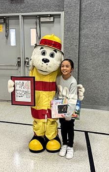 Sparky the Fire Dog costume with a young girl holding a drawing and a box, standing in a gymnasium.