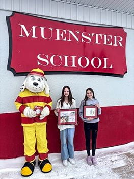 Sparky the Fire Dog with two girls holding framed pictures in front of a "MUENSTER SCHOOL" sign outside.