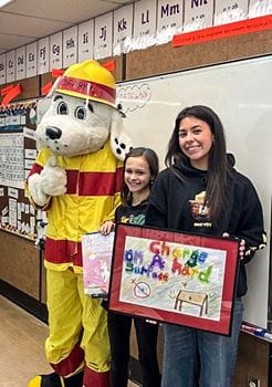 Sparky the Fire Dog Mascot posing with two girls in a classroom. One holds a prize, the other a framed picture.