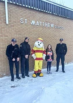 A group of five individuals, including a young girl, firefighters and Sparky the Fire Dog, stands in front of a brick wall labeled "ST MATTHEW SCHOOL" with snow on the ground.