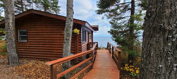A log cabin with a wooden walkway and a view of a lake, surrounded by trees.
