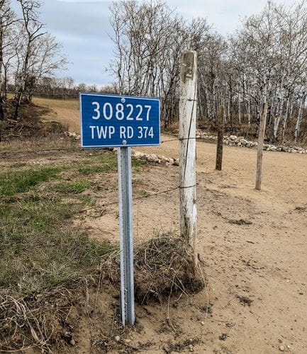 Blue rural address sign in front of an old wood fence and some trees on a dirt road